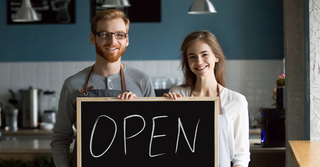 A man and woman in a cafe hold an 'open' sign