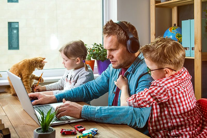A dad works whilst keeping kids and cat busy|A mum and her girl work together on a laptop|A dog rest on its owners lap as he works on a laptop|a man workas at a desk with a happy child underneath whilst a woman works behind on a sofa