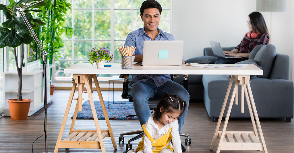 a man workas at a desk with a happy child underneath whilst a woman works behind on a sofa