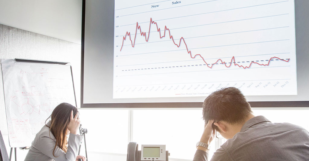 Two colleagues sit sadly with head in hands at a graph showing a slump in sales.