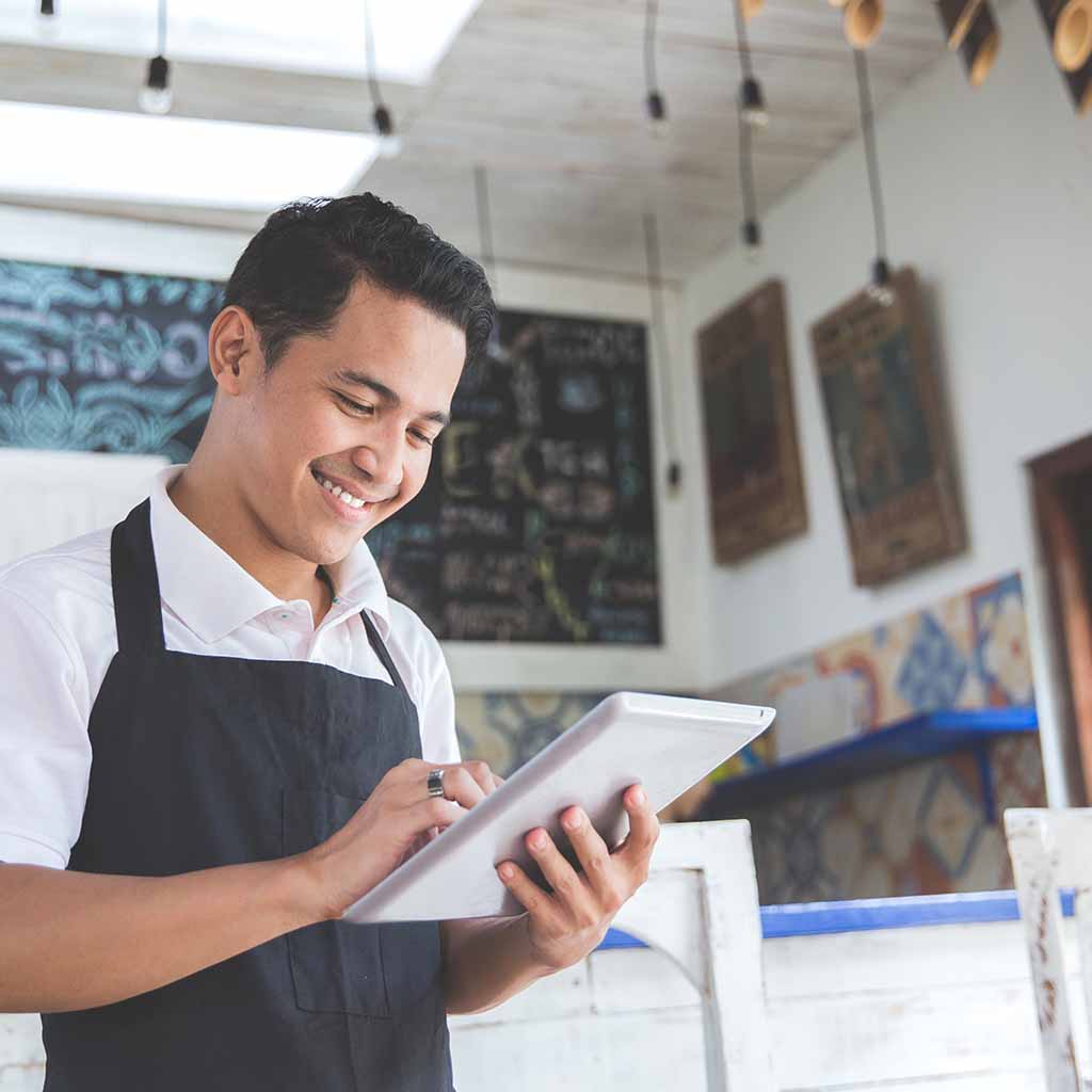 A man in an apron looks at his ipad in a coffee shop.