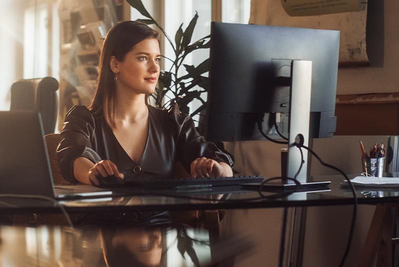 A woman sits at an office computer with sun streaming through the windows.