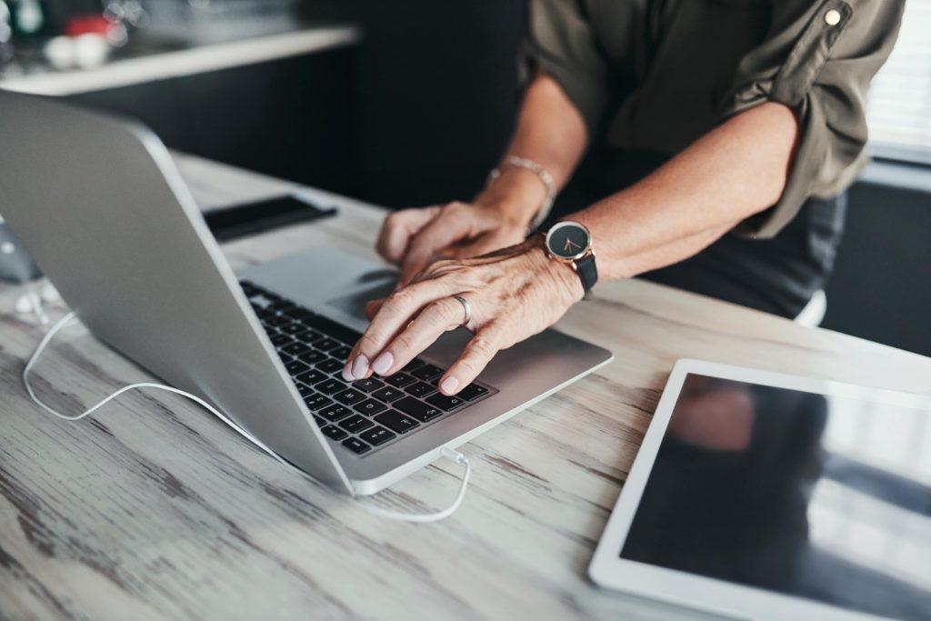 An SEO copywriter's arm with a watch and gold ring on the finger typing on a silver laptop. There's a tablet next to it on the desk.